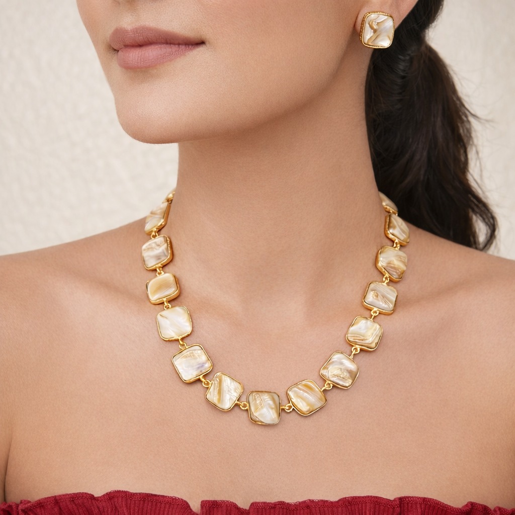 Close-up of a woman wearing a gold-tone necklace with square mother-of-pearl stones and matching stud earrings, paired with an off-shoulder red top against a soft neutral background.