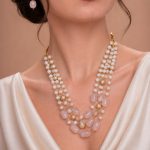 Model wearing a multi-layered gold-toned necklace featuring white pearls and blush pink oval beads, paired with matching drop earrings against a neutral background.