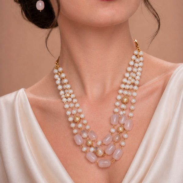 Model wearing a multi-layered gold-toned necklace featuring white pearls and blush pink oval beads, paired with matching drop earrings against a neutral background.